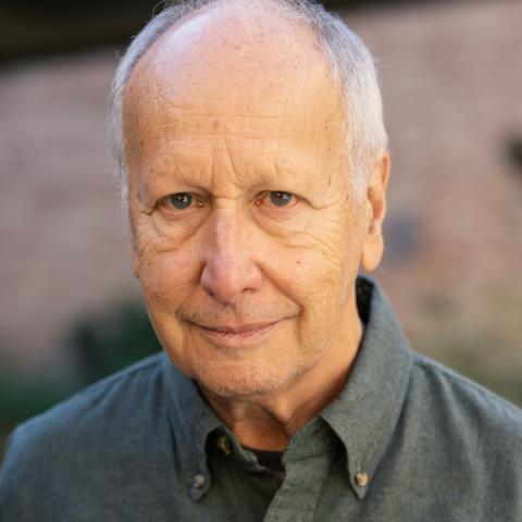 A man smiling slightly, he is dressed in a dark, green shirt. His background is blurred. 