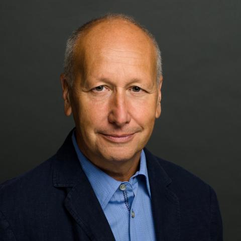 Man smiling slightly with a dark background. He is dressed in a blue shirt and dark blazer. 